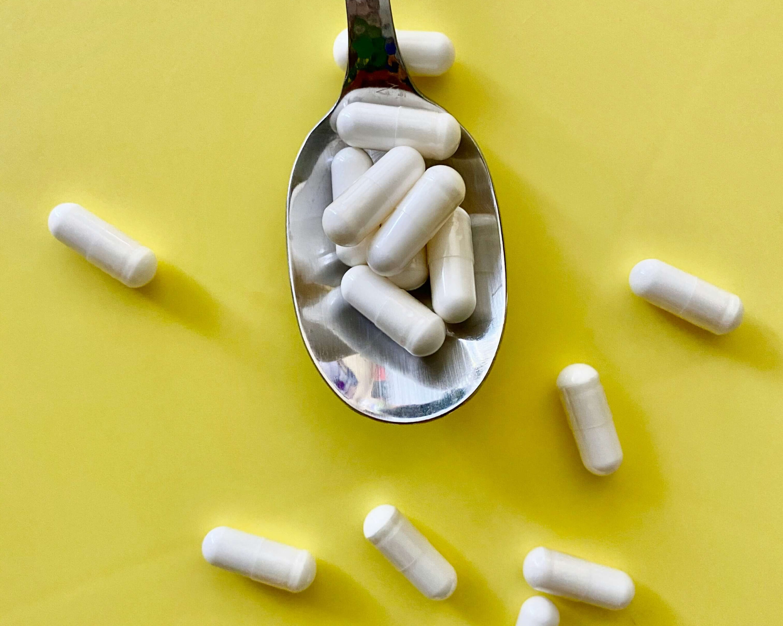 Ein silberner Löffel voller weißer Kapseln liegt auf einem leuchtend gelben Hintergrund; ringsum verstreut sind weitere Kapseln – symbolisch für postbiotische Nahrungsergänzung bei PCOS.